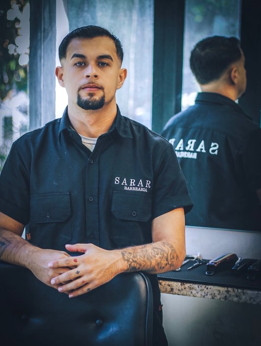 Barbeiro da Sarart sentado na cadeira, de camisa preta da casa, olhando para a câmera.
