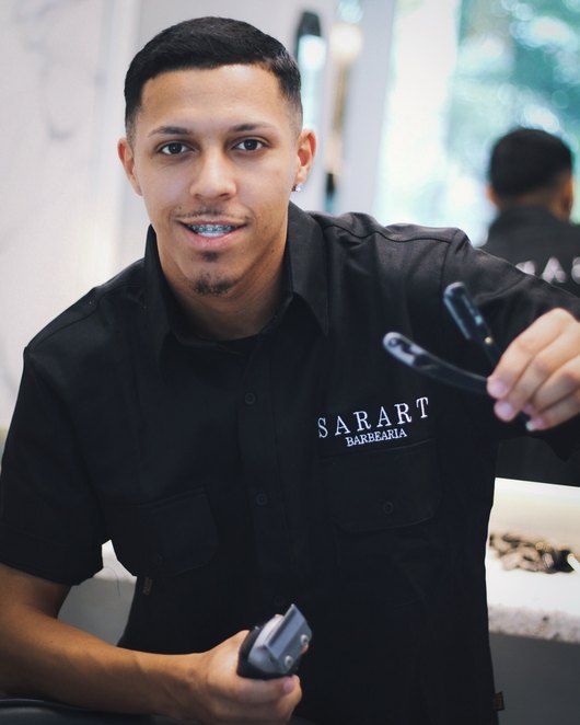 Barbeiro da Sarart sorrindo, de camisa preta da casa, com a máquina na mão.