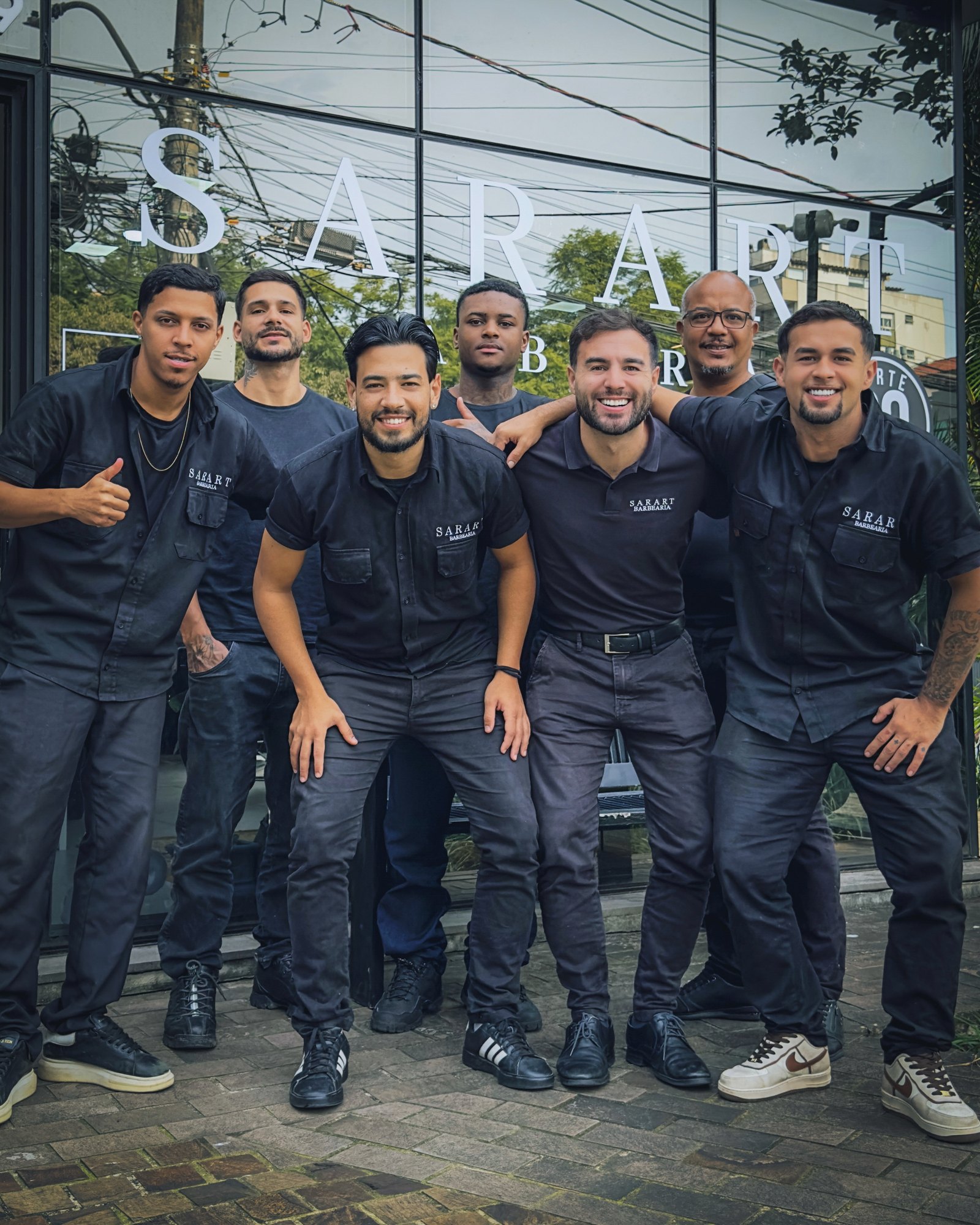 Os barbeiros da Sarart reunidos na calçada, em frente à fachada de vidro com as letras Sarart Barbearia.