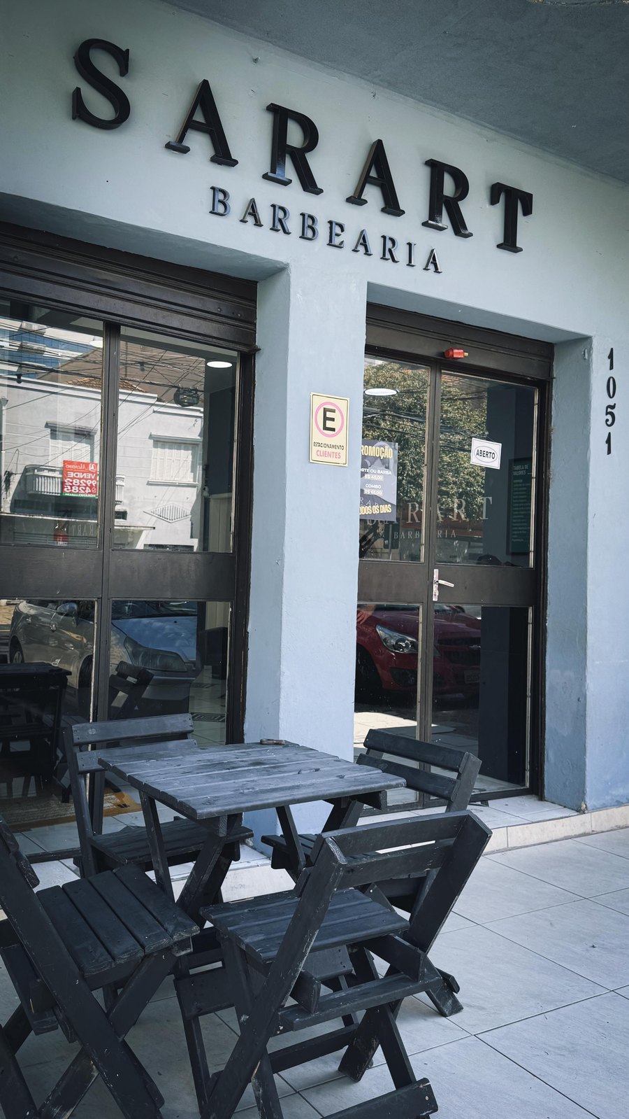 Fachada da unidade Santana: letras Sarart Barbearia sobre a porta, número 1051 na lateral e mesinhas de madeira na calçada.